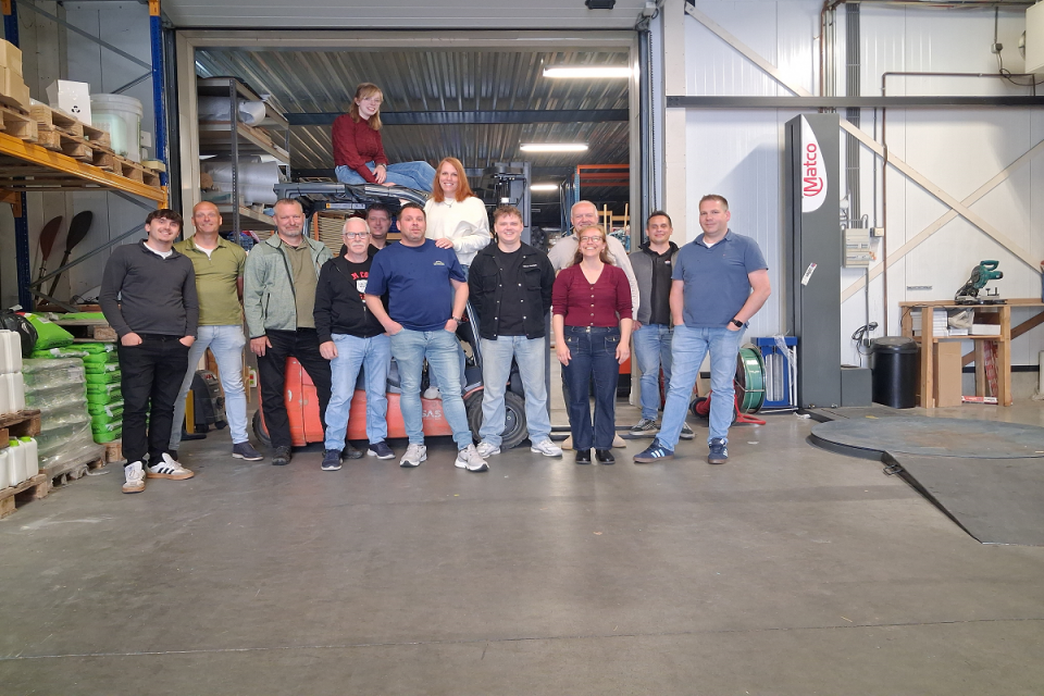 Groepsfoto van het Uniwarm-team in het magazijn, met collega&rsquo;s die samen poseren tussen stellingen, materialen en bedrijfsapparatuur.